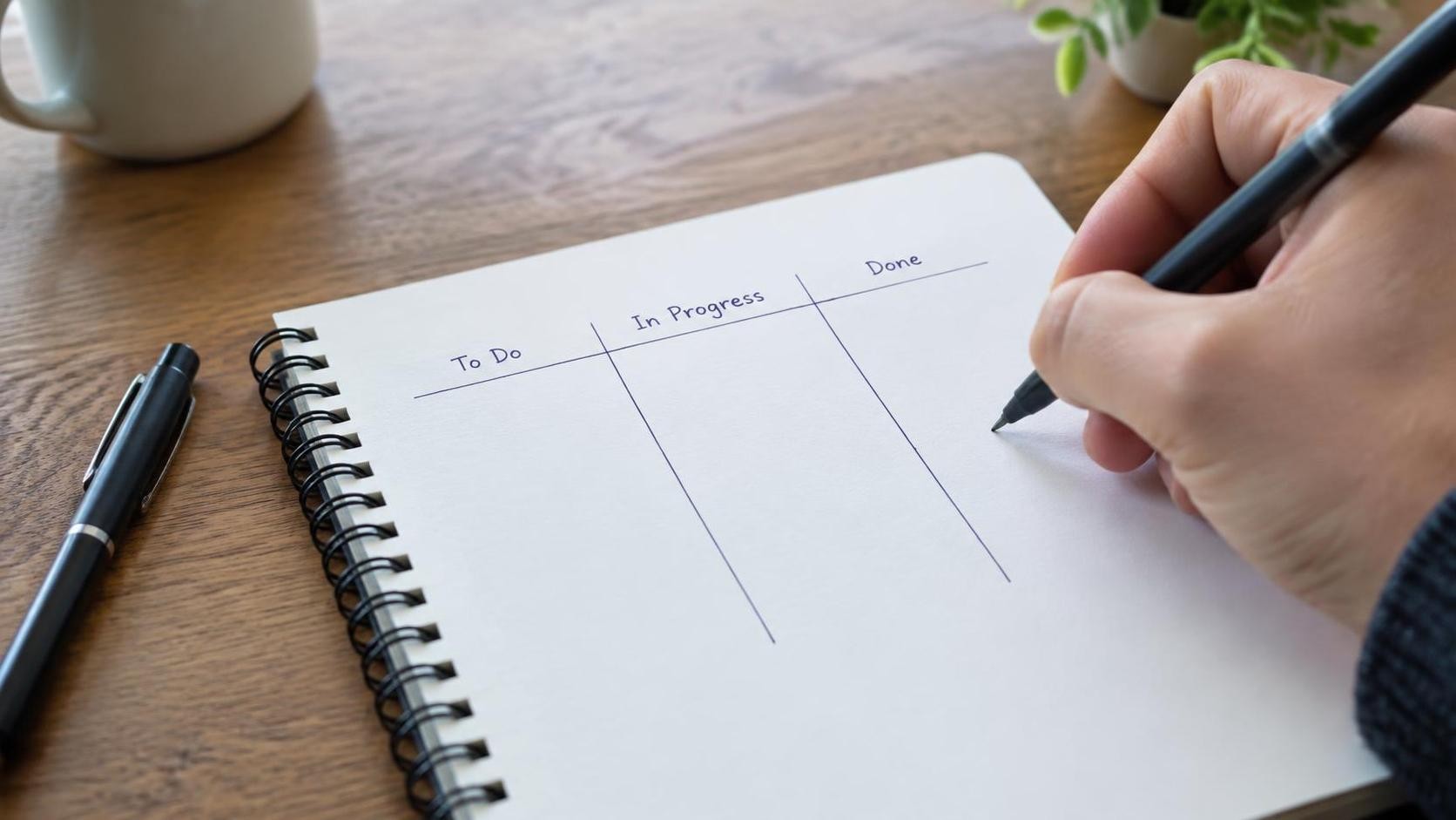 English, photorealistic, close-up of a hand drawing three labeled columns on a blank notebook page, a pen resting beside it, soft daylight on a wooden table, shallow depth of field, minimal text, 16:9